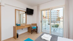 Une chambre d’hôtel comprenant un lit, un bureau avec un miroir, une chaise, un téléviseur fixé au mur et un balcon donnant sur des immeubles.