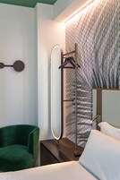 A hotel room with a green chair, a bedside table with a lamp, a large mirror, and a patterned wallpaper.