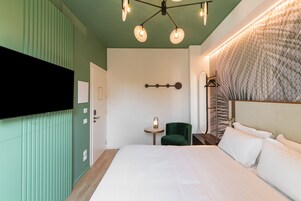 A hotel room with a large bed, a TV mounted on the wall, a small round table, and a green chair.