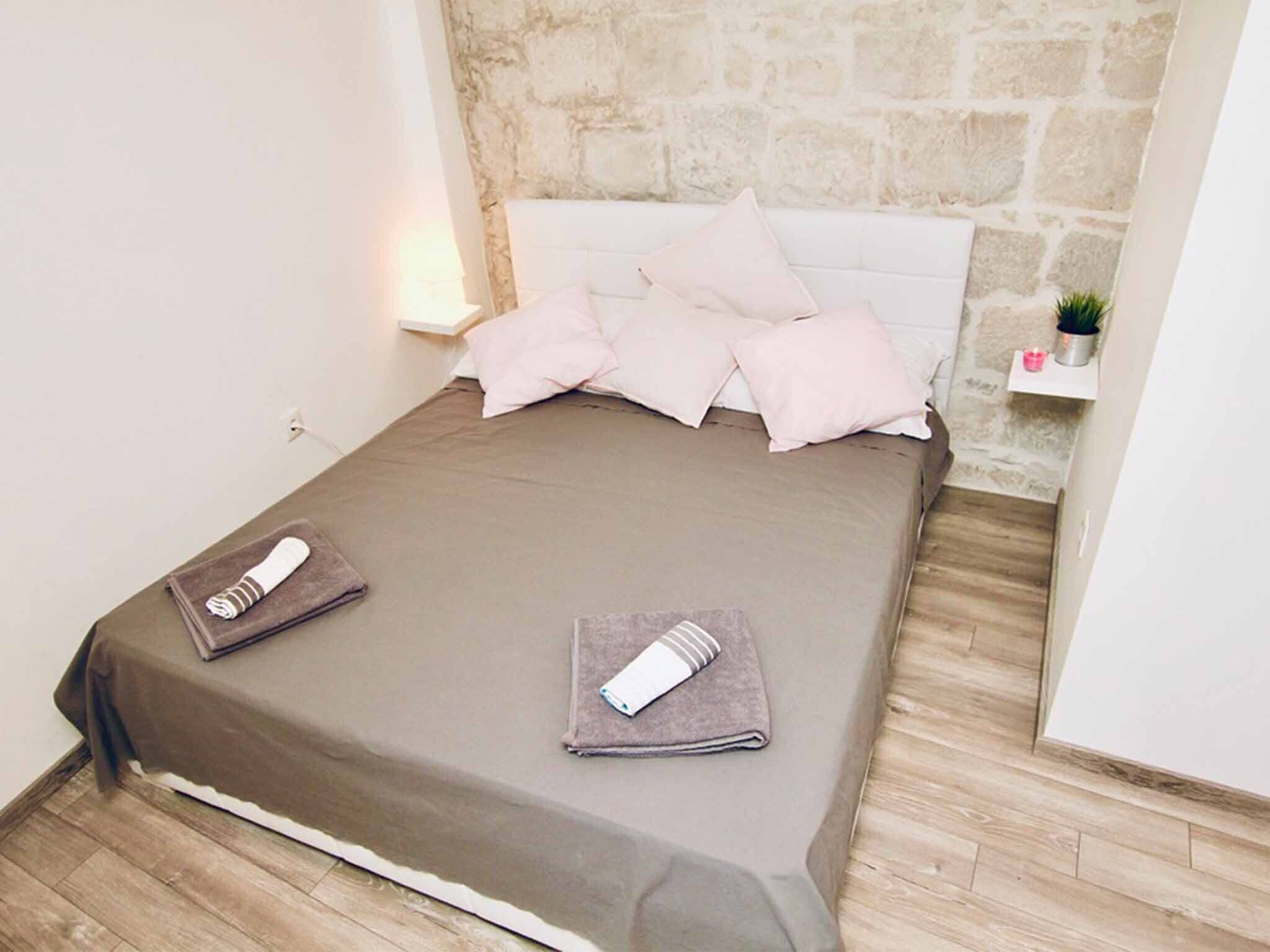 A neatly made bed with a brown bedspread and two towels, a stone wall behind, and a small shelf with a plant and candle.