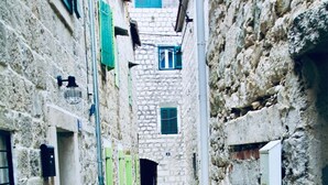 Un estrecho callejón empedrado flanqueado por edificios, con pequeñas ventanas con contraventanas y algunos tableros eléctricos.