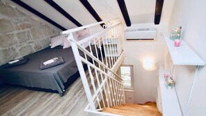 Un dormitorio con cama, escalera con barandilla blanca, pared de piedra y una ventana pequeña.