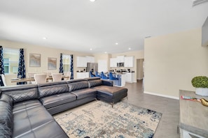 A modern living room with a sectional sofa, a dining area with a table and chairs, and a kitchen with white cabinets and appliances. - 7655 Oakmoss Loop (Davenport)