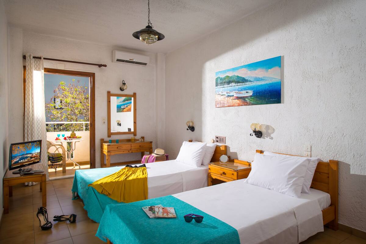 A hotel room with two beds, a TV, a desk, and a balcony with a view of the sea.