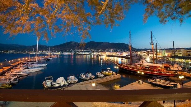 A marina at dusk with boats docked, a large ship, and a cityscape in the background. - Floral Hotel (Hersonissos)