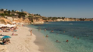 A beach with clear water, people swimming and sunbathing, and buildings on a hillside in the background. - Floral Hotel (Hersonissos)