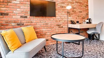 A room with a brick wall, a flat-screen TV mounted above a wooden table, a grey sofa with yellow cushions, and a patterned carpet.