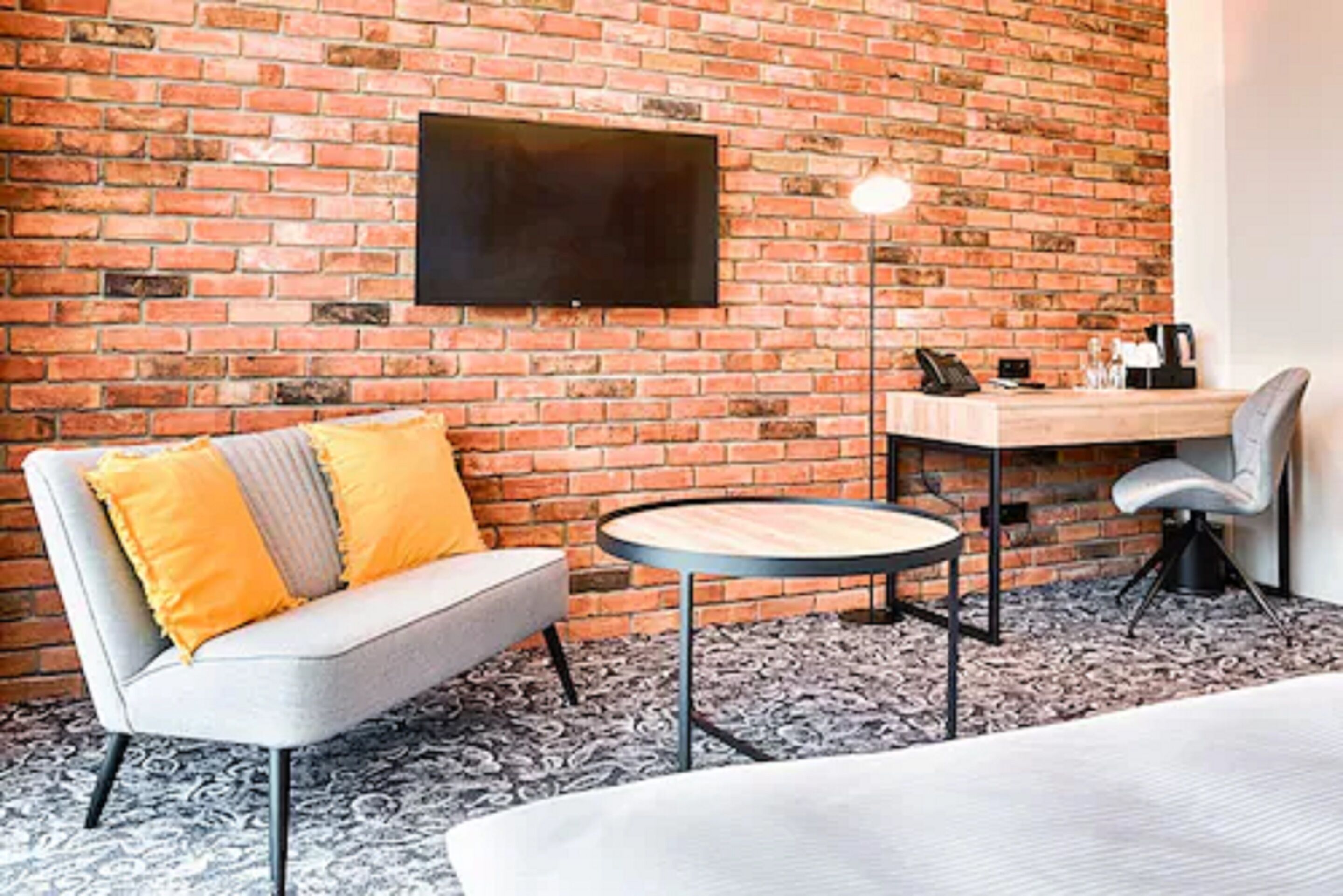 A room with a brick wall, a flat-screen TV mounted above a wooden desk, a grey sofa with yellow cushions, a round table, and a chair.