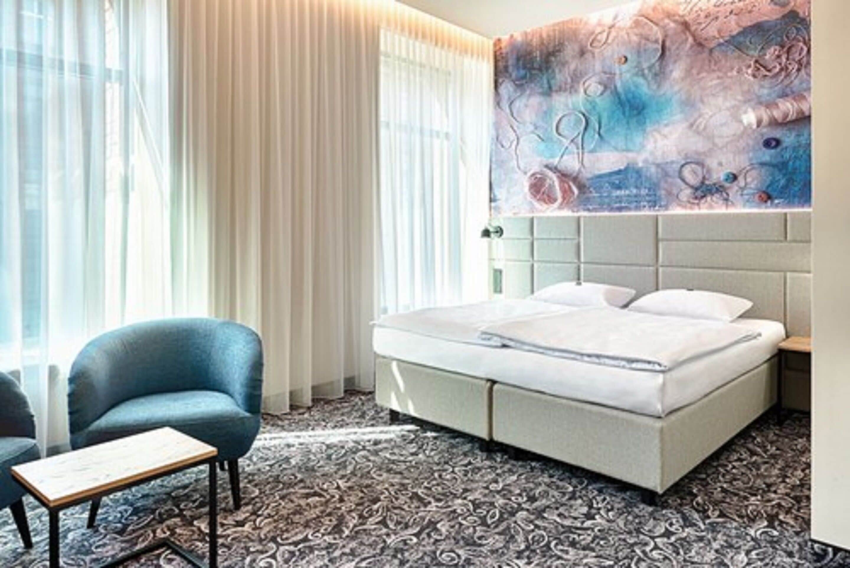 A hotel room with a bed, a chair, a small table, and a large abstract painting on the wall.