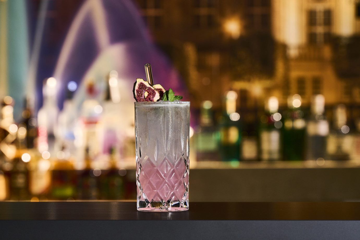 A cocktail glass with a pink hue, garnished with a slice of fruit and mint, placed on a bar counter with a blurred background of bottles and a cityscape.