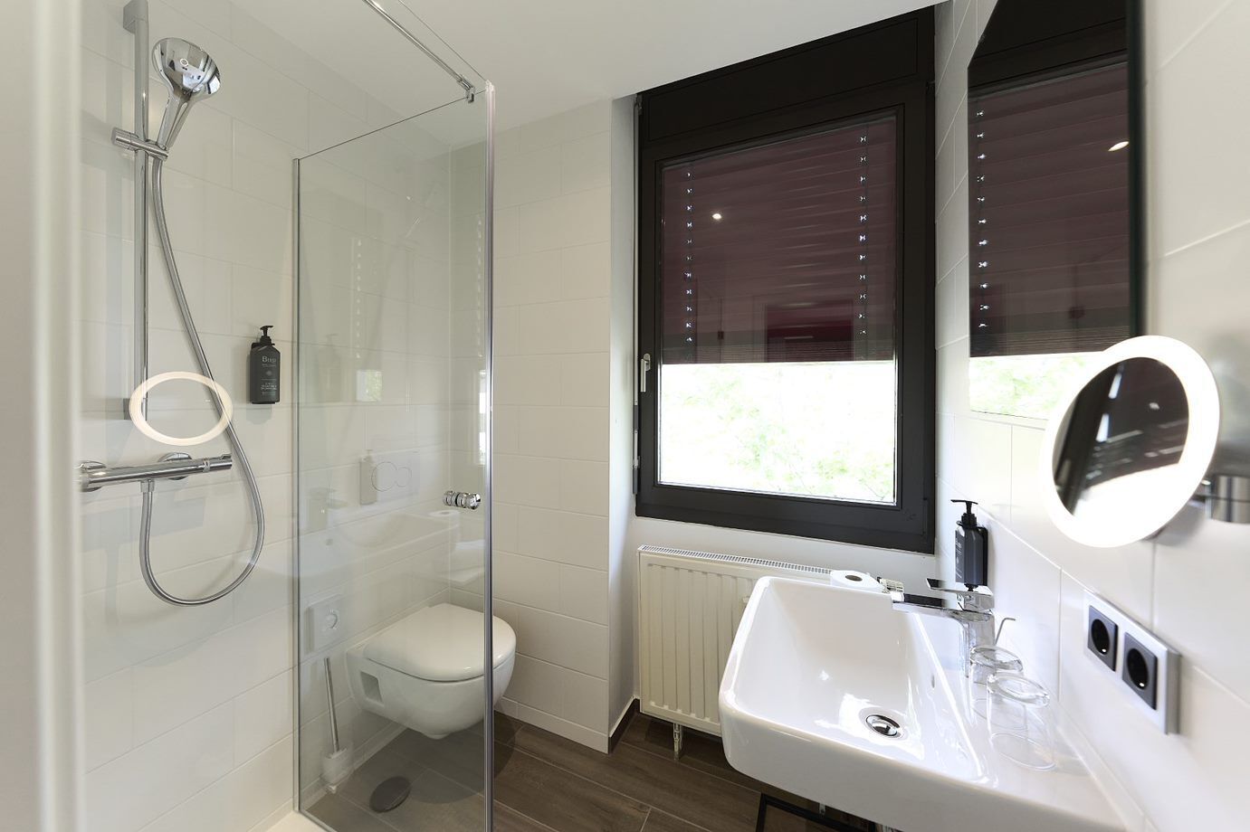 Un baño moderno con ducha envuelta en vidrio, un lavabo blanco y un ventanal con persianas.