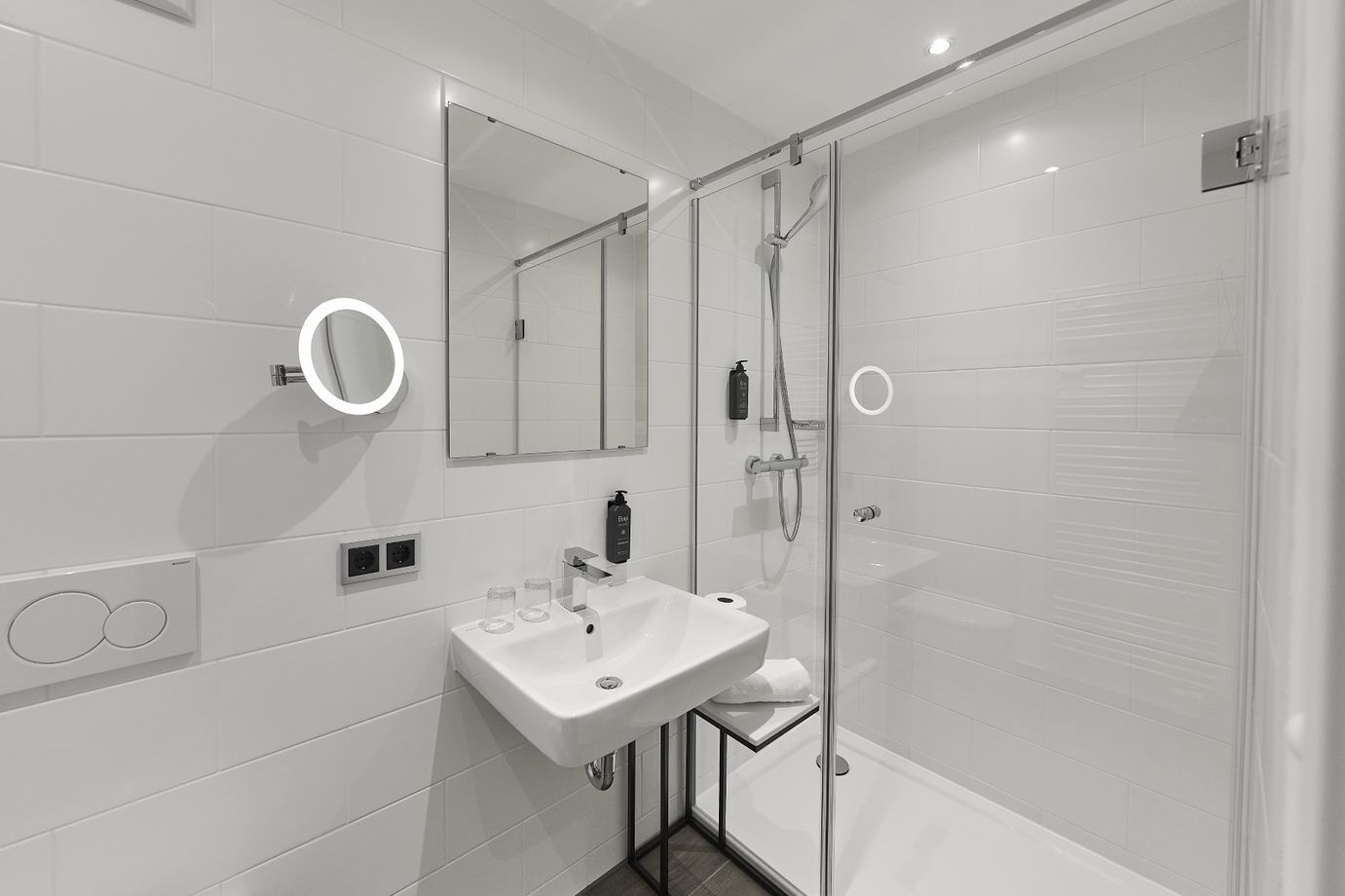 Een moderne badkamer met een douche in een glazen cabine, een wit wastafelmeubel met een spiegel erboven en een ronde wandlamp.
