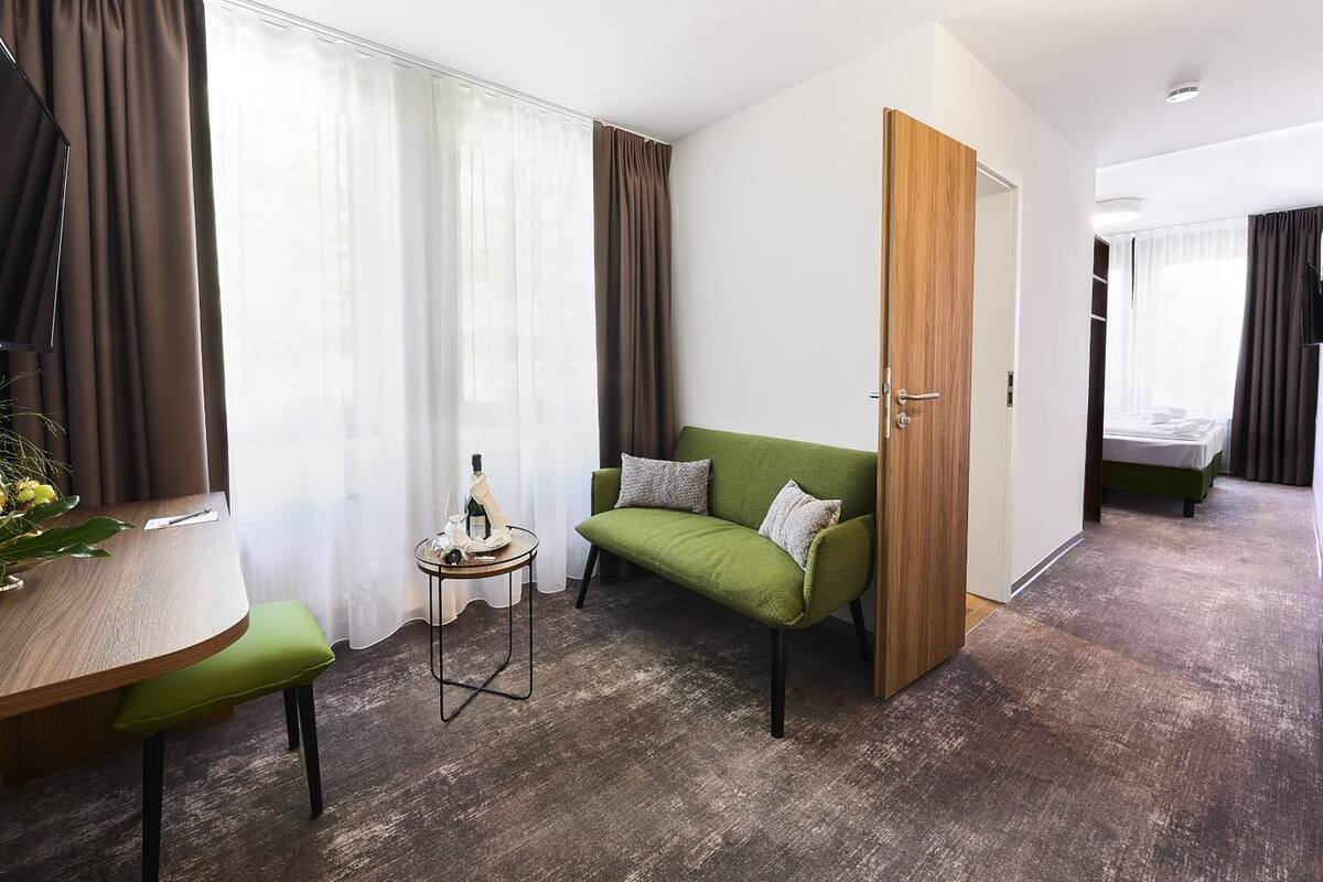 A modern hotel room with a green sofa, a small round table with a bottle and glasses, a wooden door, and a bed in the background.