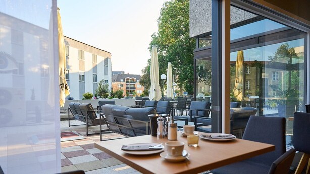Un espace extérieur aménagé avec des tables et des chaises, de grandes fenêtres et une vue sur les bâtiments et les arbres.