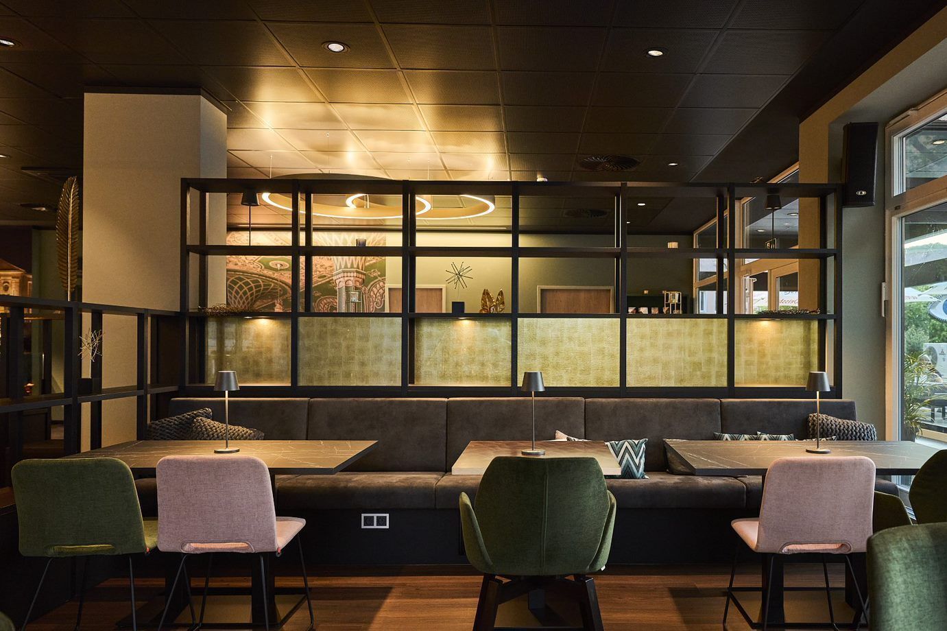 Un restaurante moderno con techo alto, iluminación empotrada y una zona de estar con sillas tapizadas en verde y un banco acolchado.