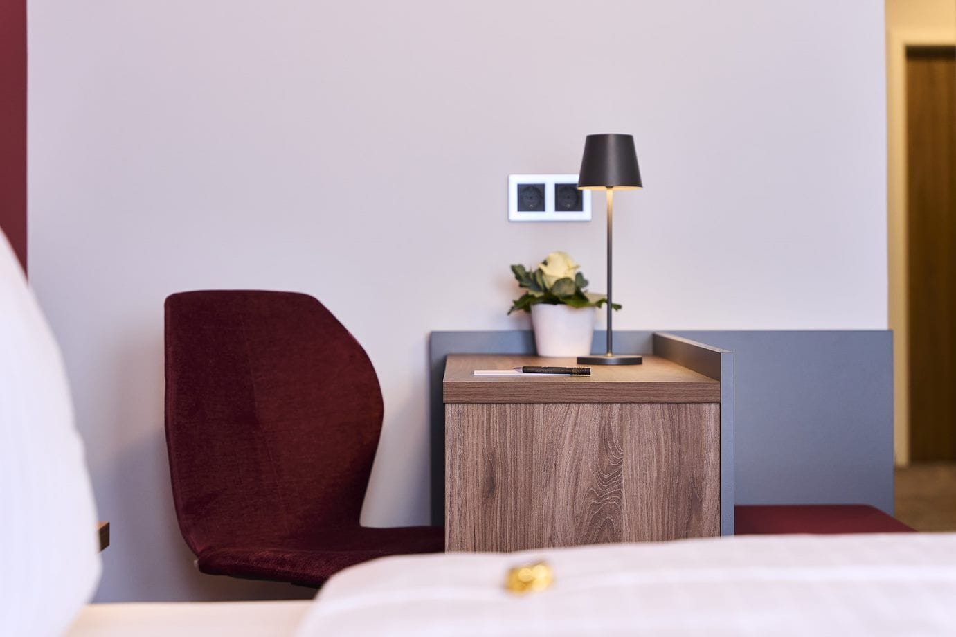 Une chambre d’hôtel avec un fauteuil rouge, une petite table d’appoint en bois sur laquelle se trouve une lampe, une plante en pot et un lit avec une literie blanche.