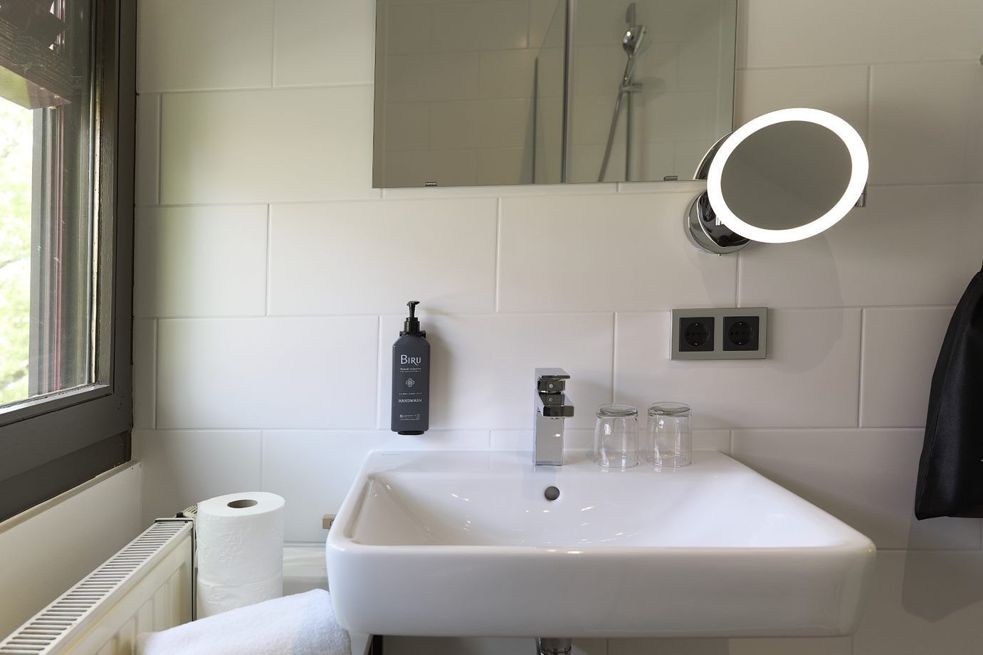 Ein Badezimmer mit einem weißen Waschbecken, einer Handseifen-Spender, einem Spiegel mit Beleuchtung und einer sichtbaren Dusche im Hintergrund.