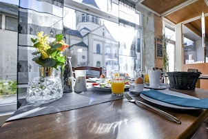 Una mesa con desayuno: un florero con flores, una jarrilla, un vaso de jugo de naranja y un plato con huevos y tostadas.