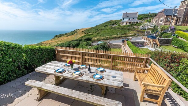 A wooden deck with a picnic table set for four, overlooking a coastal landscape with houses and greenery. - Rosemary Cottage - Three Bedroom House, Sleeps 5 (Mortehoe)