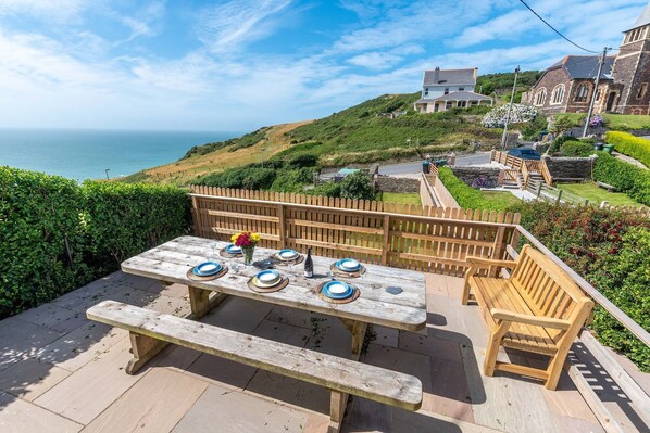 Eine Holzterrasse mit einem Picknicktisch für vier Personen, mit Blick auf eine Küstenlandschaft mit Häusern und viel Grün.