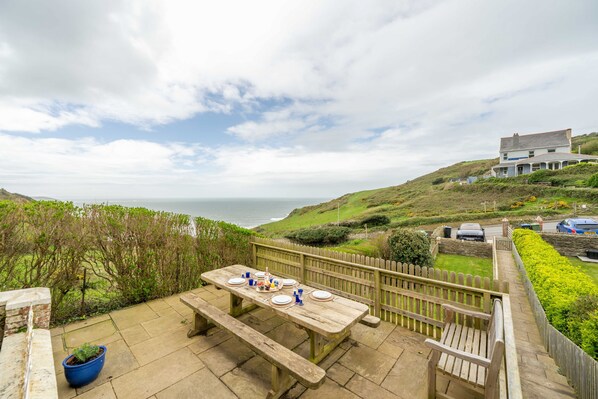 A wooden deck with a table set for dining, overlooking a coastal landscape with a house and parked cars. - Rosemary Cottage - Three Bedroom House, Sleeps 5 (Mortehoe)