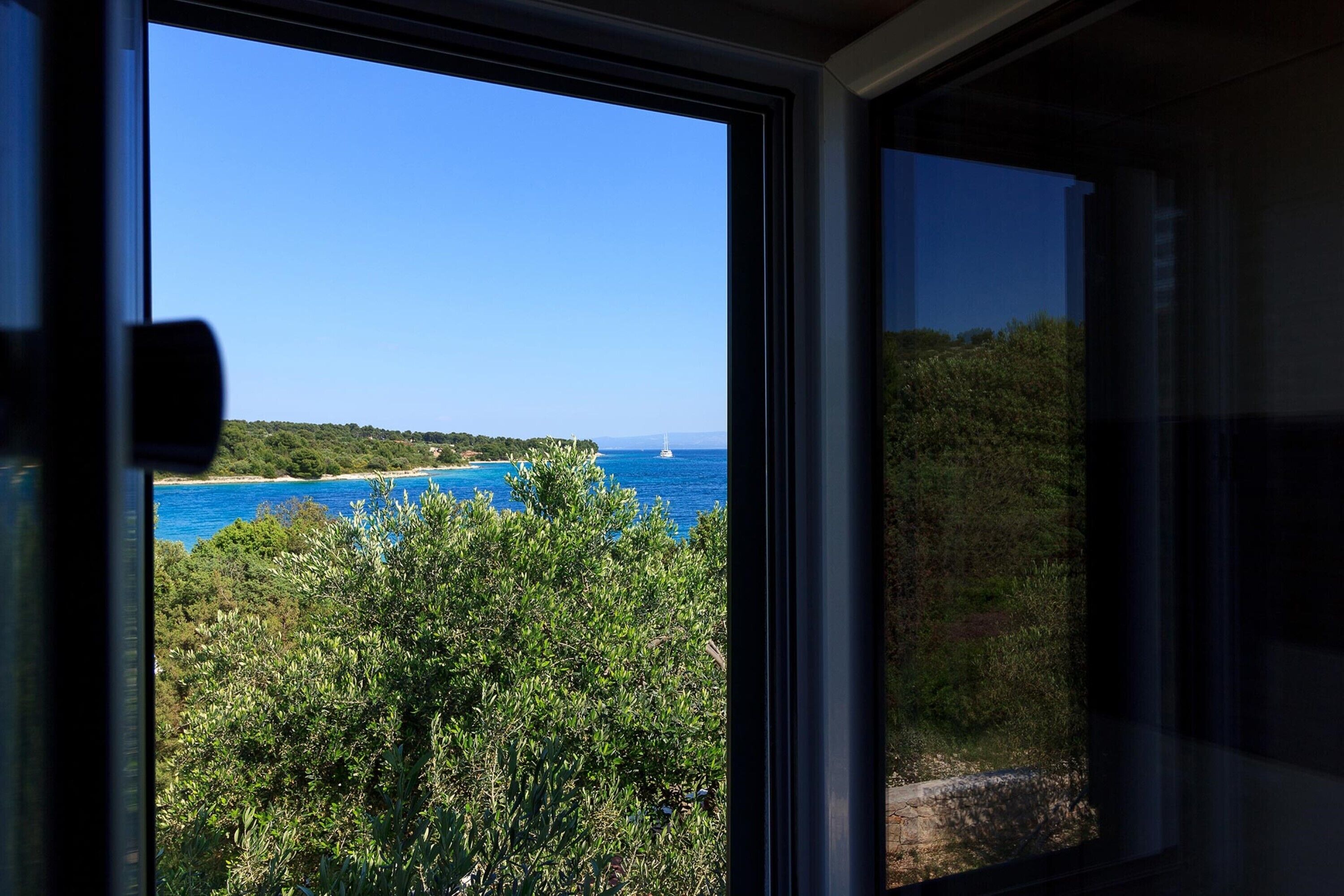 En vy genom ett fönster som visar ett kustlandskap med klar himmel, tät vegetation och en vattenyta.