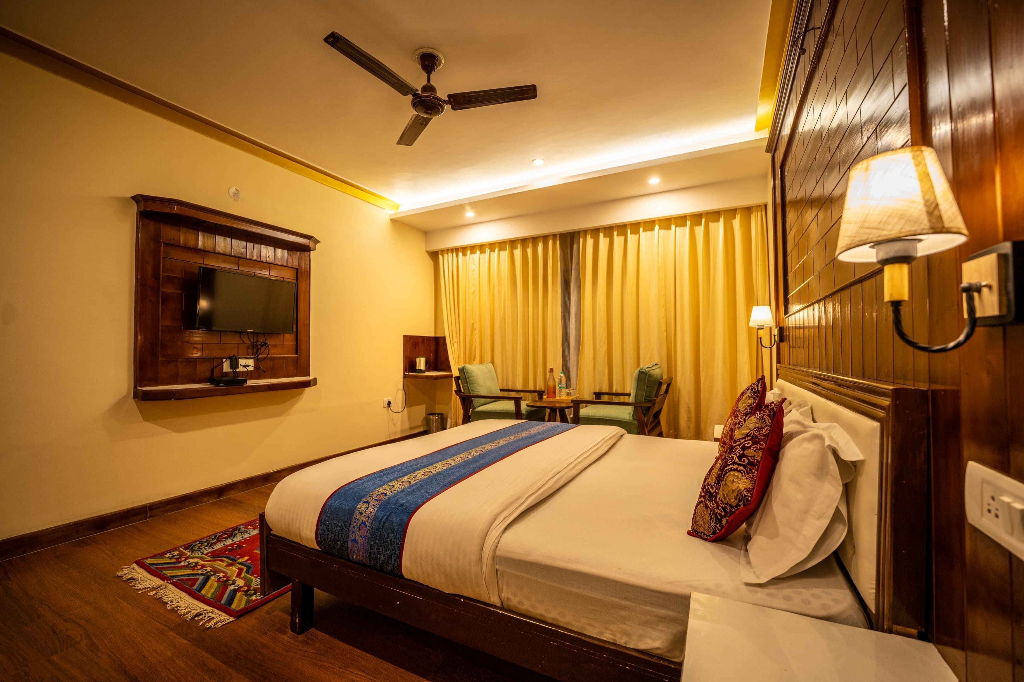 A hotel room with a bed, a television mounted on the wall, a ceiling fan, a table with chairs, and a lamp.