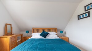 Un dormitorio con una cama de madera, una colcha azul acolchada, dos mesitas de noche con lámparas, un aparador de madera y un espejo.
