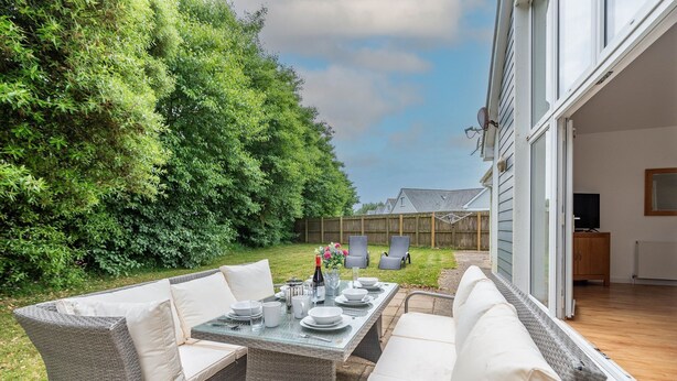 Un patio con mobiliario de mimbre y una mesa de vidrio lista para comer, rodeado de exuberante follaje y vistas a una casa.