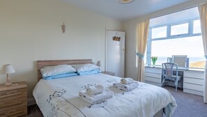 Una cama bien hecha con un edredón floral, una mesita de noche de madera con una lámpara, una silla junto a la ventana y vista al mar.