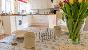 Una cocina con armarios blancos, una lavadora, una mesa con mantel estampado con corazones y un florero con tulipanes.