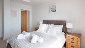 A neatly made bed with white linens and rolled towels, a wooden headboard, and a bedside table with a lamp and decorative item.