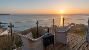 A balcony with wicker chairs and a table, overlooking a sunset over the ocean.