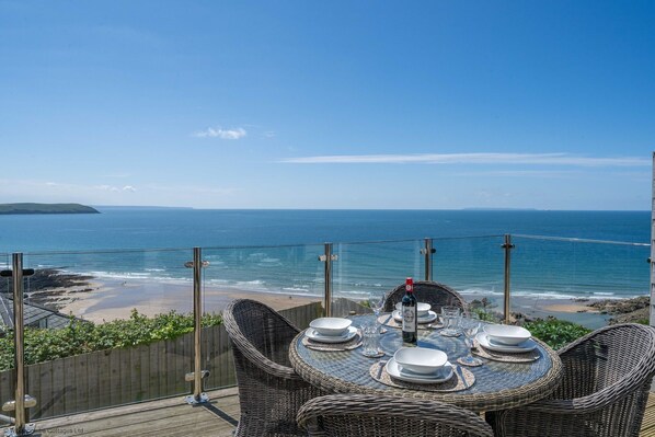 A dining table set with white plates and glasses, overlooking a beach and ocean.