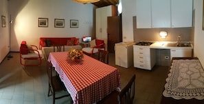 Cocina compacta con estufa a gas, lavaplatos y armarios blancos. Hay una zona de comedor con mantel a cuadros rojo y blanco y un jarrón de flores.