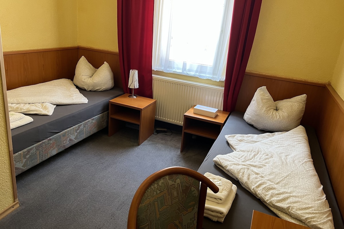 A compact hotel room with two single beds, a small wooden nightstand, a radiator, and a window with red curtains.