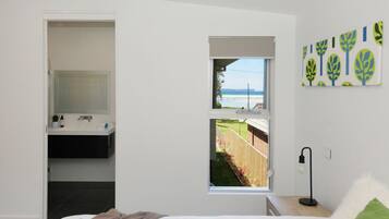 A modern bathroom with a large mirror, a sink, and a shower. There is a window with a view of the beach and a fence.