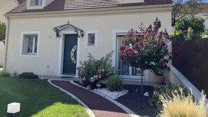 Ein zweistöckiges Haus mit einem dunkelbraunen Dach, einer schwarzen Haustür und einer kleinen Veranda mit weißer Brüstung. Blumen und ein Weg führen zur Tür.