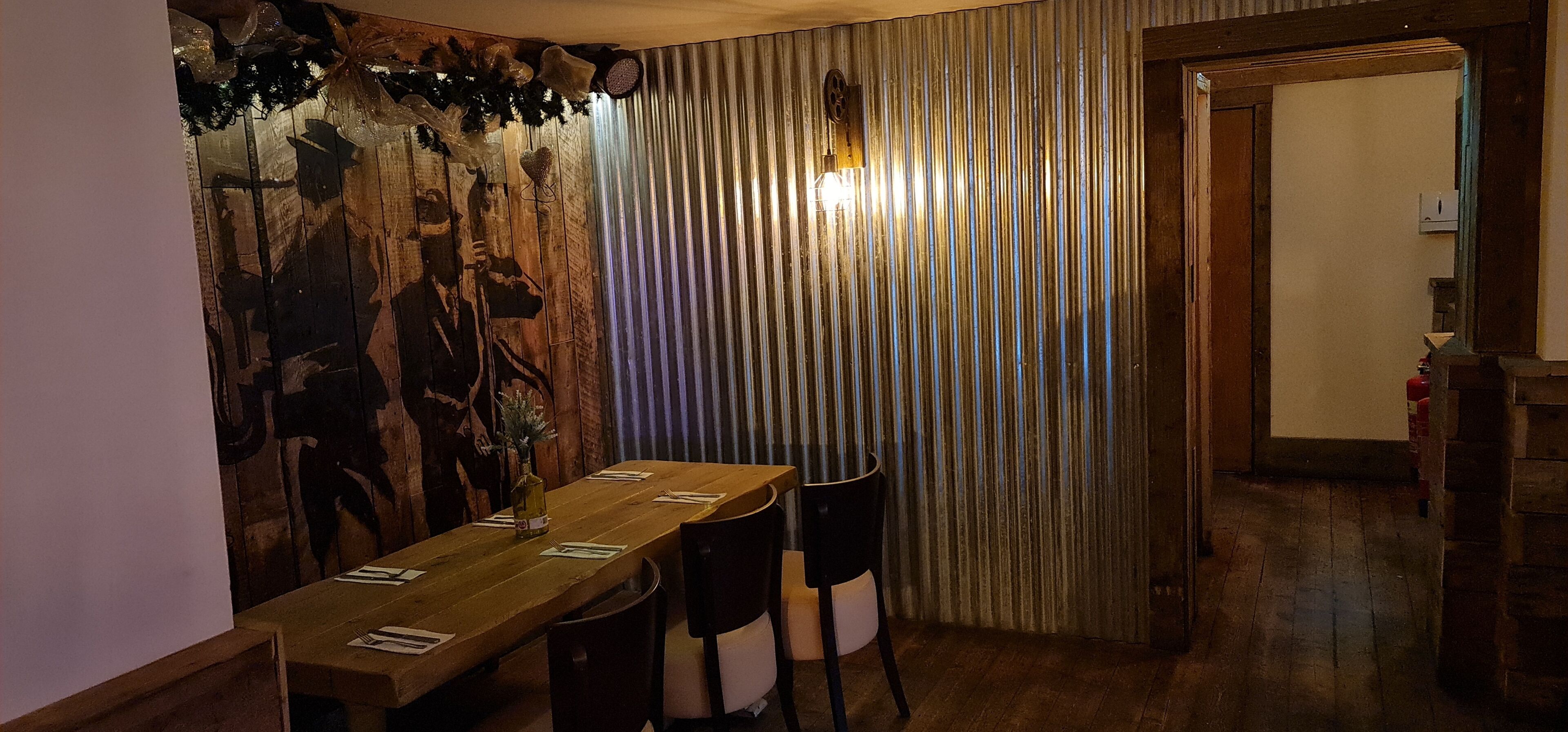 Un comedor con una larga mesa de madera dispuesta para dos comensales, una pared decorativa con diseño rústico y una cortina a rayas al fondo.