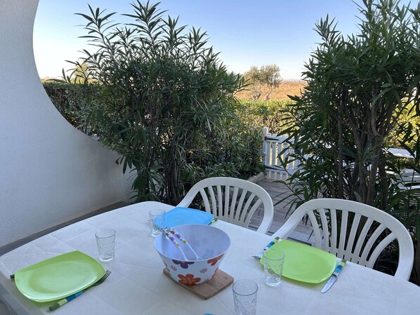 Ett bord dukat utomhus med vita stolar, en vit duk och utsikt över en trädgård med höga växter och ett vitt staket i bakgrunden.