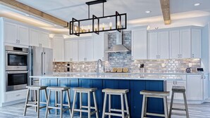 A modern kitchen with white cabinets, a blue island with bar stools, stainless steel appliances, and a brick backsplash. - Brand New Construction with a Private Pool, Water Views, & Parking for Four! (Cape May)