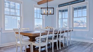 A dining area with a wooden table and white chairs, large windows, and a hanging light fixture. - Brand New Construction with a Private Pool, Water Views, & Parking for Four! (Cape May)