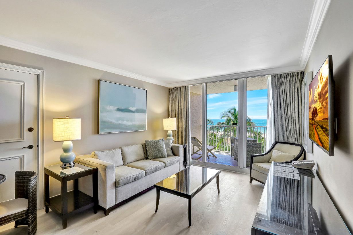 A modern hotel room with a sofa, a coffee table, a TV, and a balcony with a view of the ocean.
