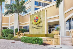 The exterior of Marco Beach Ocean Resort with a prominent crest sign, a hotel restaurant sign, and a well-maintained landscaped area with palm trees and shrubs. - Lavish Beachfront Condo | Rooftop Pool/Spa/Gym (Marco Island)