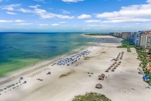 A beach with clear blue waters, a sandy shore, and a row of buildings along the coastline. - Lavish Beachfront Condo | Rooftop Pool/Spa/Gym (Marco Island)