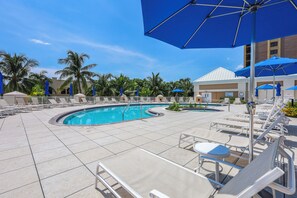 A pool area with lounge chairs and umbrellas, surrounded by palm trees and a building with a white roof. - Lavish Beachfront Condo | Rooftop Pool/Spa/Gym (Marco Island)