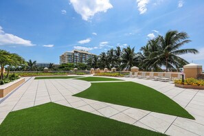 A landscaped outdoor area with artificial grass, palm trees, and a building in the background.