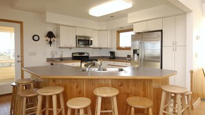 A modern kitchen with white cabinets, stainless steel appliances, a central island with stools, and a window providing natural light. - 2060 - DoodleBug - Corolla Retreat | Private Pool & Walk to Beach (Corolla)