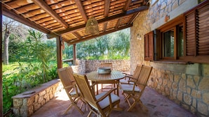 A stone patio with a wooden table and chairs, a hanging light fixture, and a stone wall with windows.