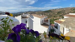 A view of white buildings with terracotta roofs, surrounded by greenery and hills in the background.